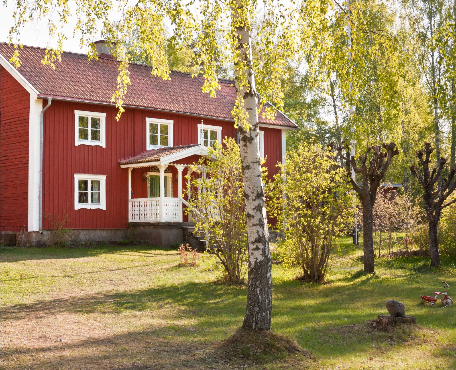 Rött trähus med vita fönsterfoder och vitt räcke vid verandan, björk i förgrunden